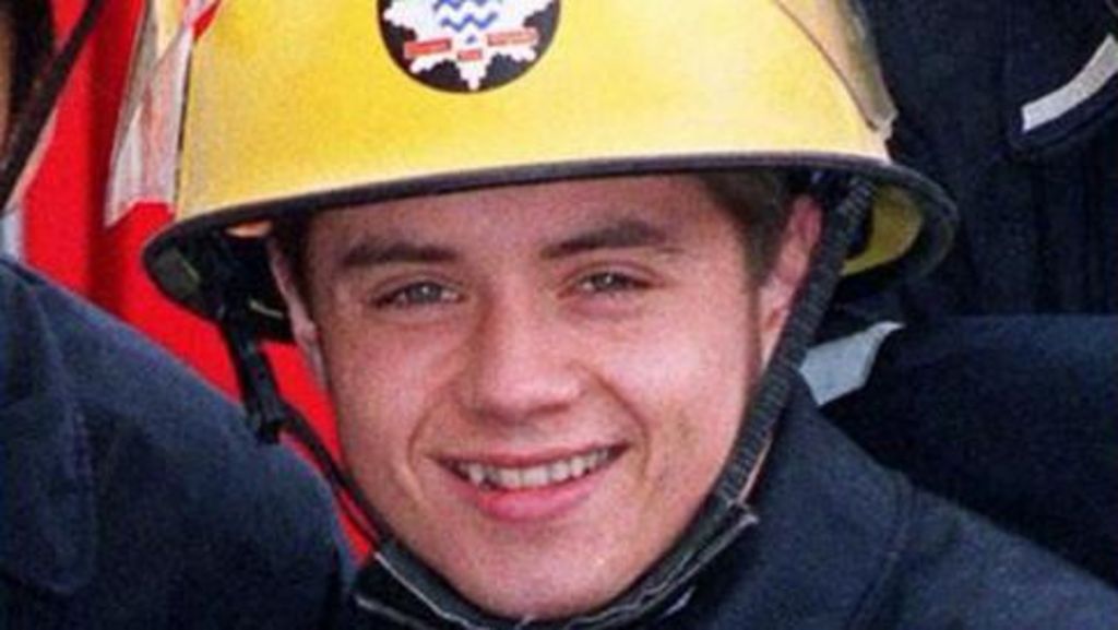 John Alford playing the part of a firefighter. He is wearing  a yellow helmet with a star-shaped badge and is smiling.
