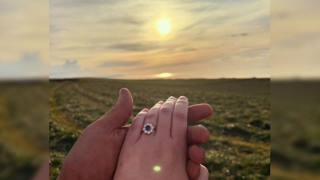 Chloe Morley and Tom Heal holding hands in the field in front of the sunset. She has a round blue engagement ring on her finger