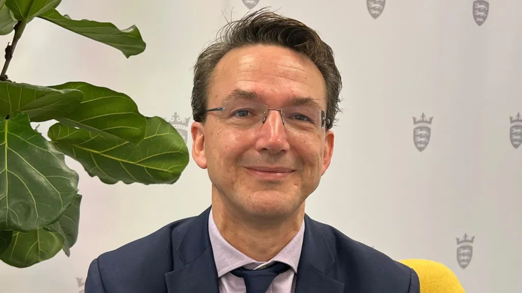 A man smiling sat on a yellow chair in front of a plant on the left and a white background with the government emblem on it.