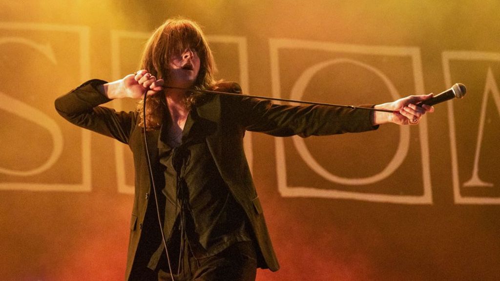 Tom Ogden on stage with his hair over his eyes and one arm horizontally outstretched and holding the microphone away from him, at The Big Feastival 2023