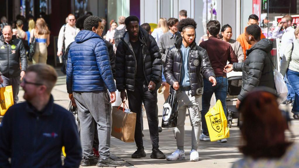A group of four young men on a busy Market Street in Manchester holding their shopping bags. Lots of other people are walking past in the background.