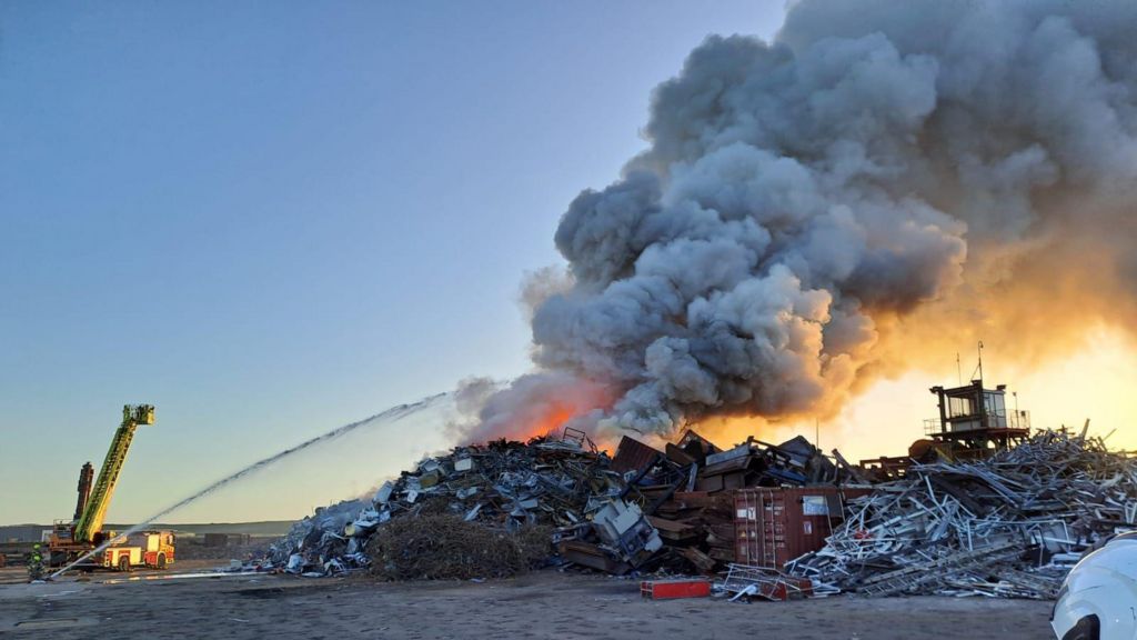 Plumes of smoke bellowing from a huge pile of scrap metal at dawn. Picture also shows a firefighter spraying a long stream of water onto the blaze from a distance