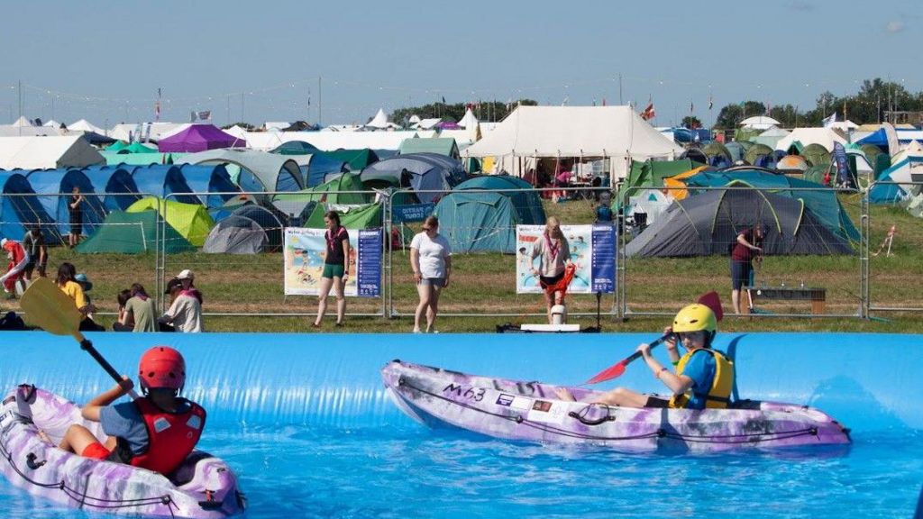 Two children canoe in a huge pool at the jamboree, with tents in the background