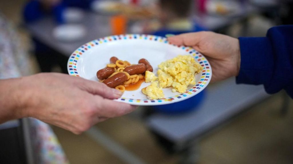 Close up of an adult hand passing a plate to a child.  The plate has some scrambled eggs, small sausage and spaghetti hoops on it.