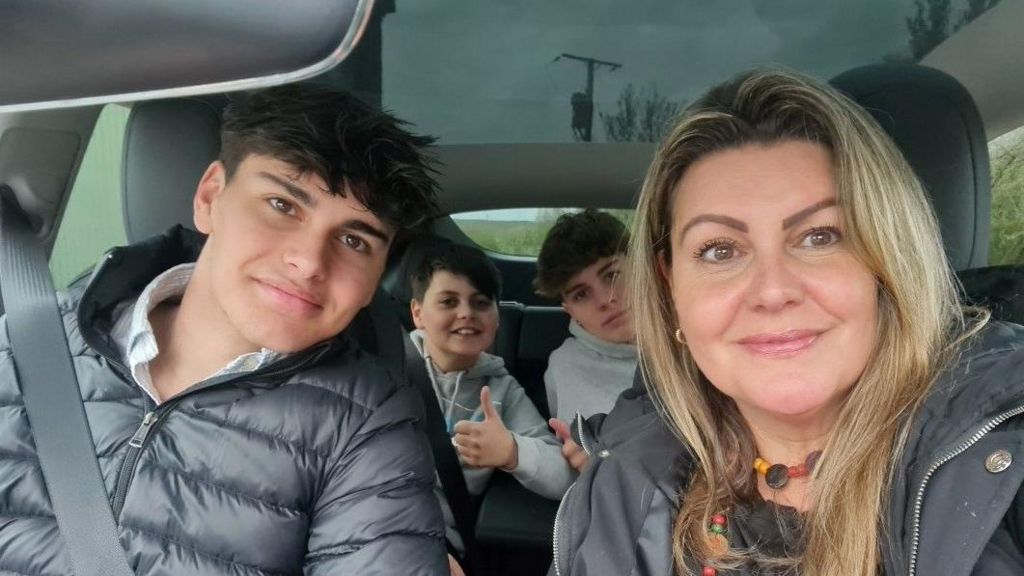 Val Brittain and her three sons smile at the camera in a selfie taken in a car, one of her sons who is sat in the back is giving the camera a thumbs up