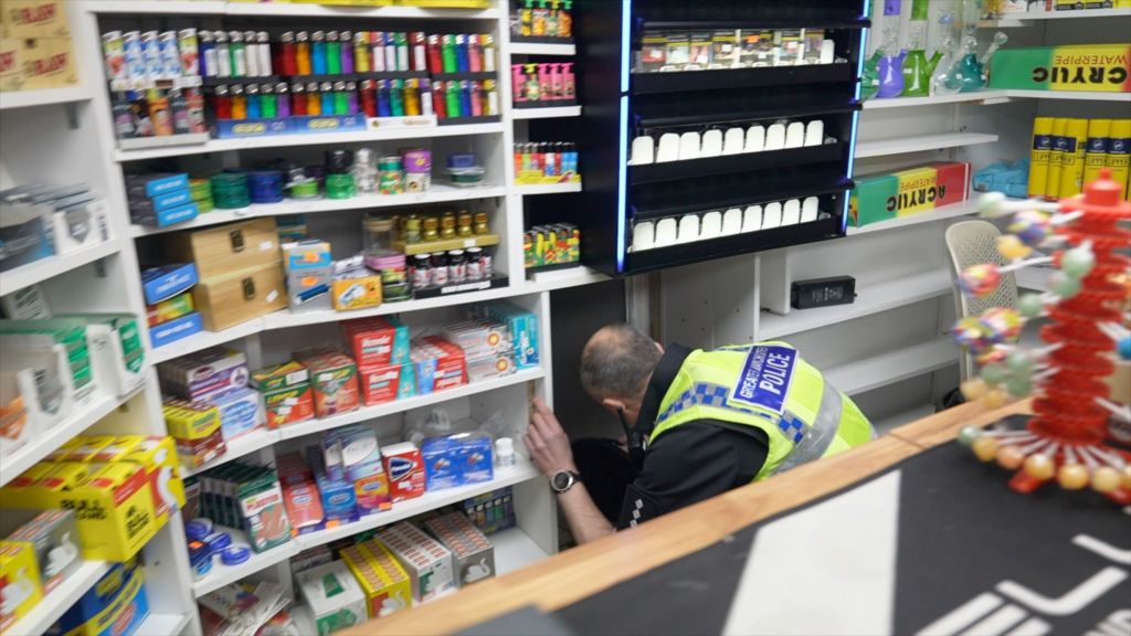 A police officer crouching down behind the counter of a mini-mart where there is a hidden compartment in the shelving, which he has opened and is looking into.