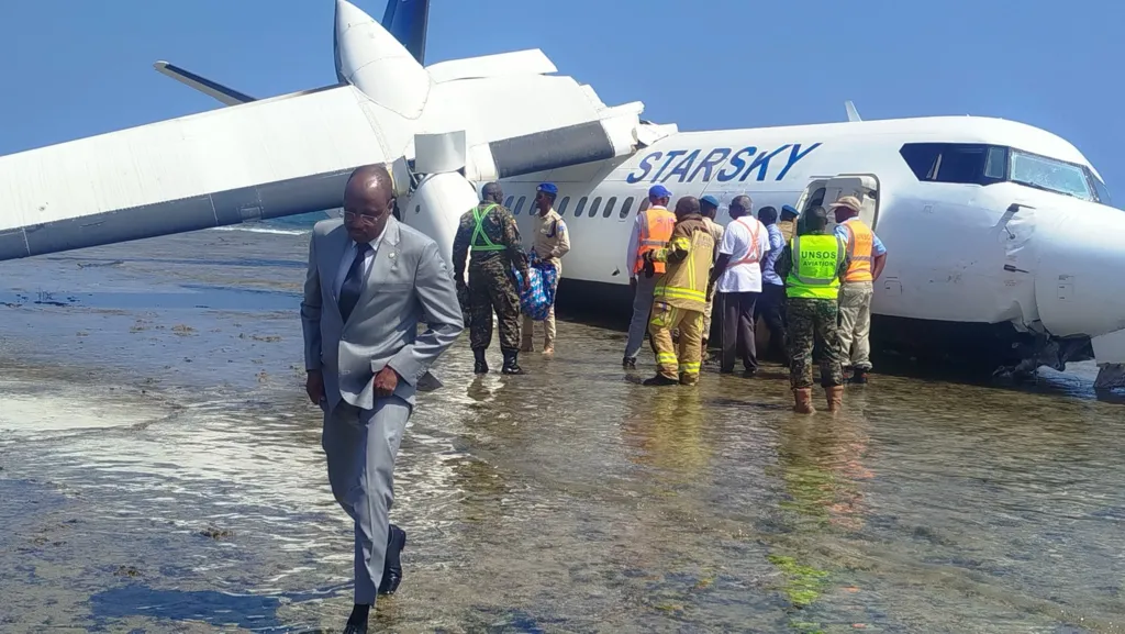 A crashed plane on the seashore is leaning over with its right wing broken. Rescue workers are close to the body of the plane and a man in a grey suit is wading through the water towards the camera.
