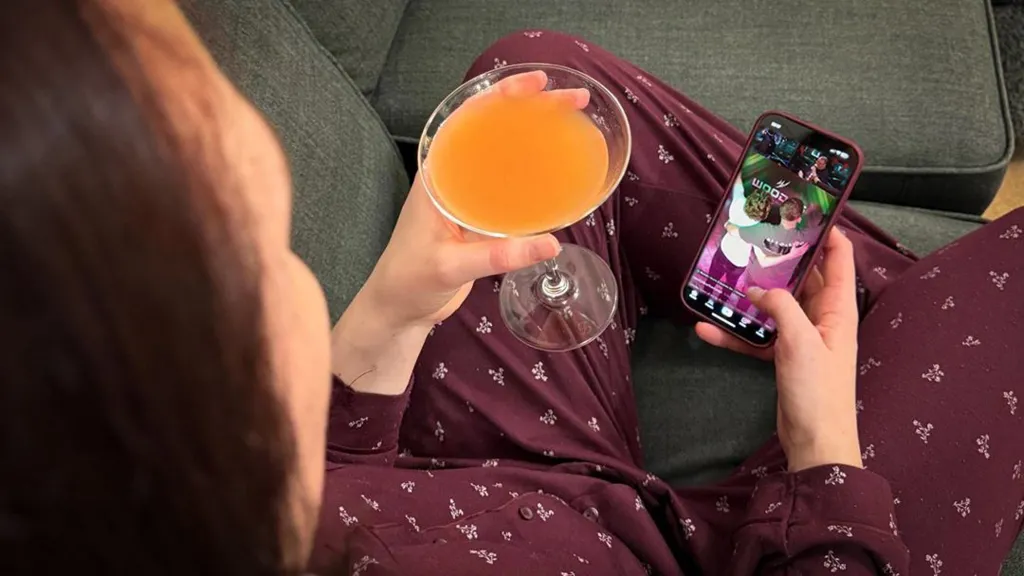 Woman sat on a sofa holding a cocktail in one hand and her phone in the other. She is watching a live stream from a bar in Manchester.  (Stream image has been added to phone by Photoshop for illustration purposes)