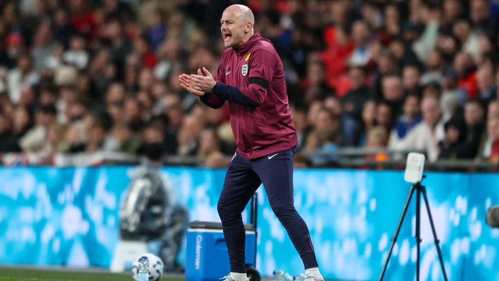 Lee Carsley standing on the side of a football pitch, with his hands in front of him. He is bald and is wearing a maroon red England jacket, with navy blue tracksuit bottoms. His mouth is open as he shouts.