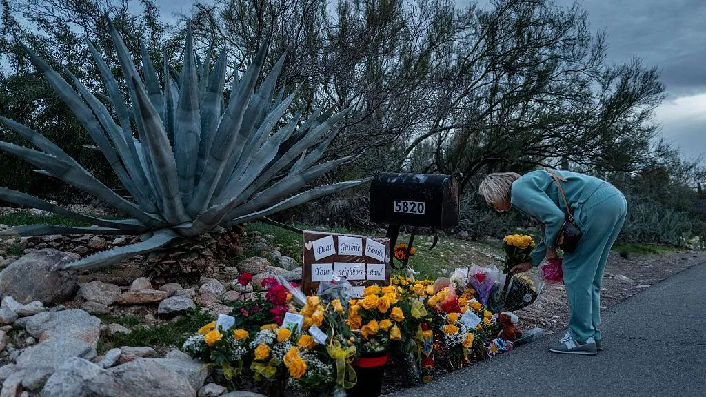 A woman in a teal sweatsuit bends over and lays flowers at a memorial for Nancy Guthrie 