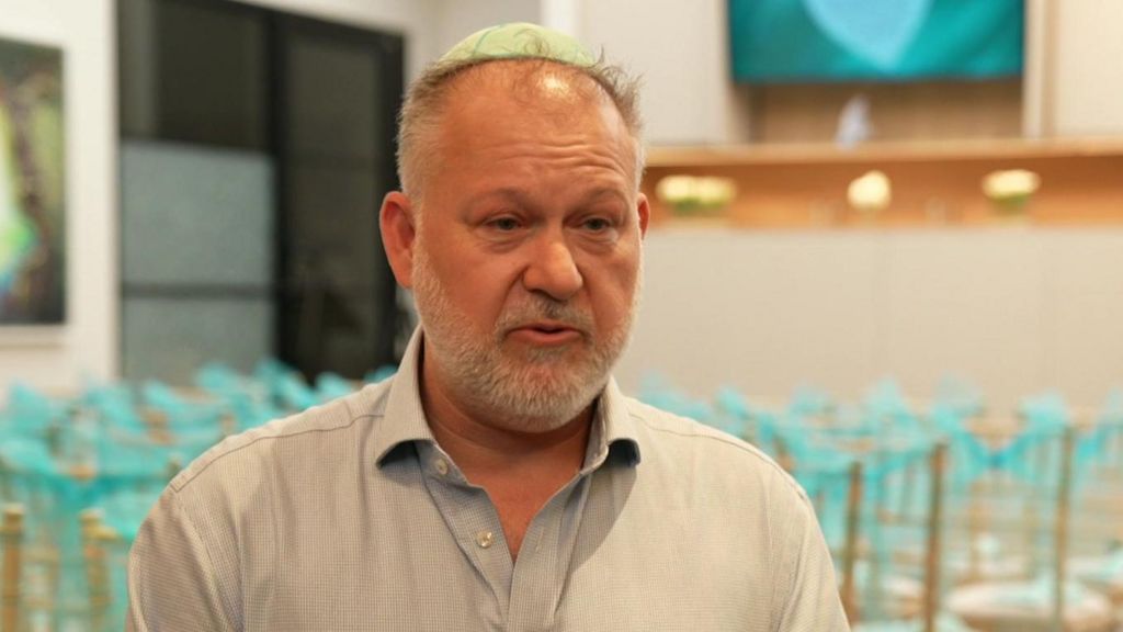 Adam Ziff wears a light green kippah and speaks while looking slightly to the side in a room with chairs covered in blue ribbons.