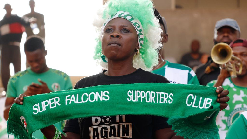 A fan of Nigeria's Super Falcons wears a green and white wig and holds up a scarf while a fellow supporter plays a trumpet in the background
