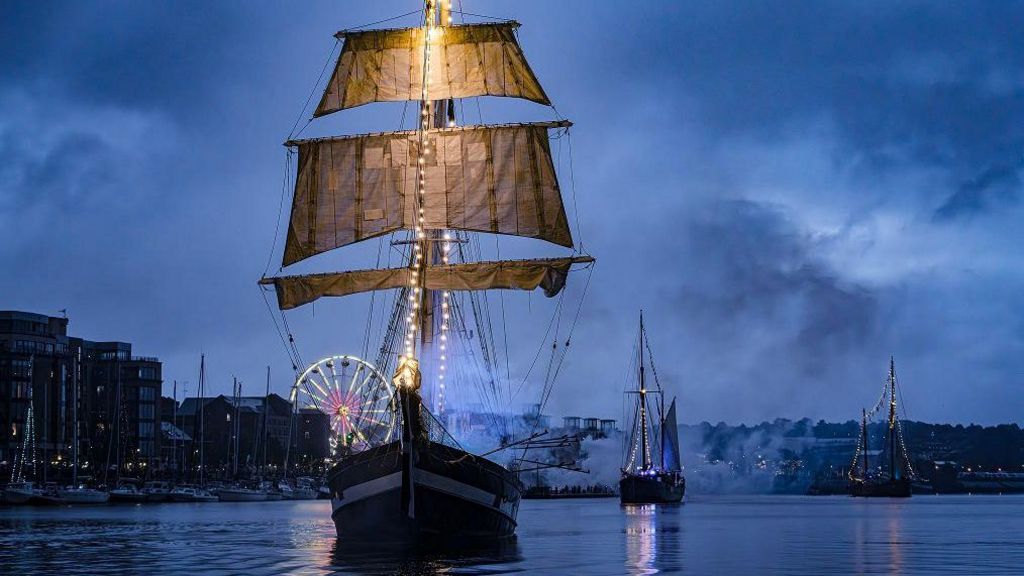 Three tall ships sailing on the Foyle river at dusk. The buildings along the quay are on the right, with an illuminated ferris wheel in the background 
