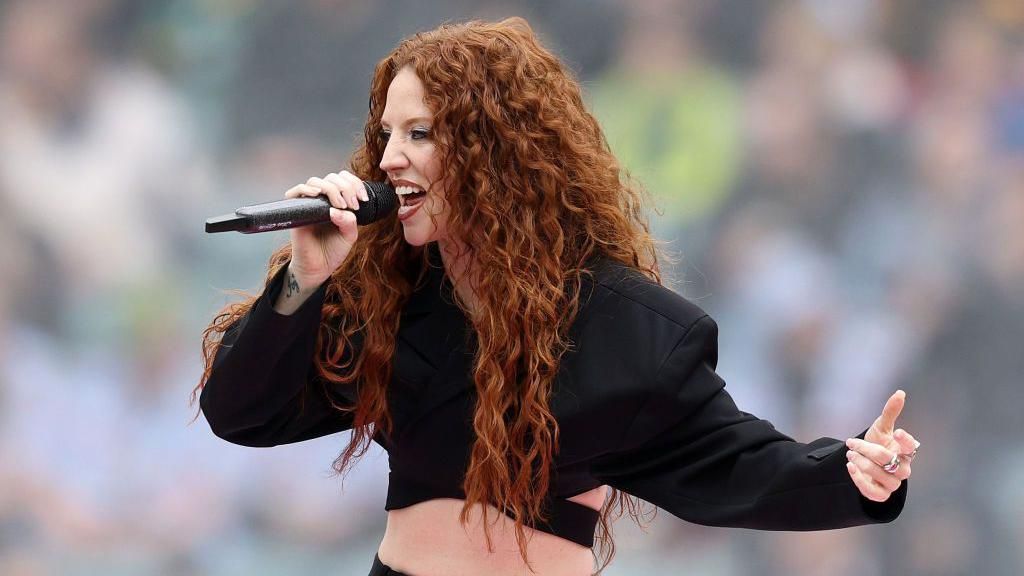 Jess Glynne is singing into a mic, she is wearing a cropped black suit jacket. SHe has long curly ginger hair. 