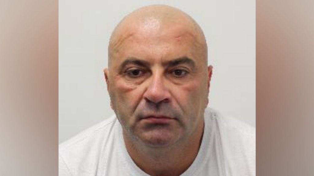 Mugshot of Bald man in white shirt looks below eyeline of camera