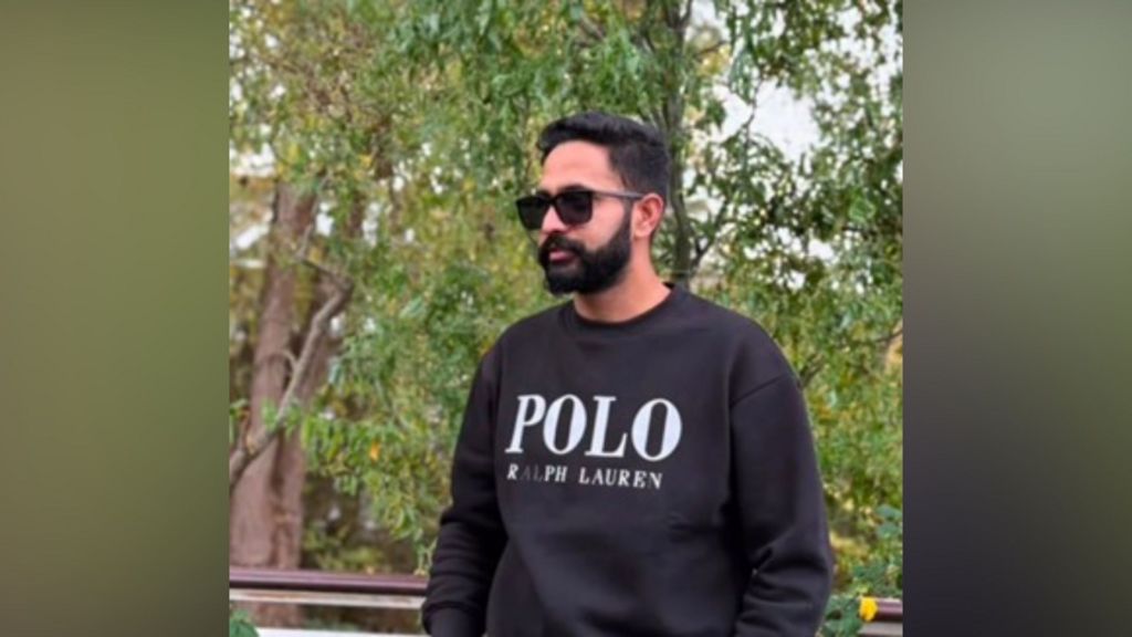 A young man standing outdoors near some trees. He is wearing a dark jumper and sunglasses and has a black beard and black hair.