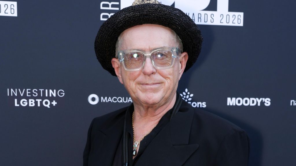 Holly Johnson on the red carpet, pictured here from the shoulders up. He is wearing a dark jacket, a chain around his neck, glasses with clear frames, and a hat. 