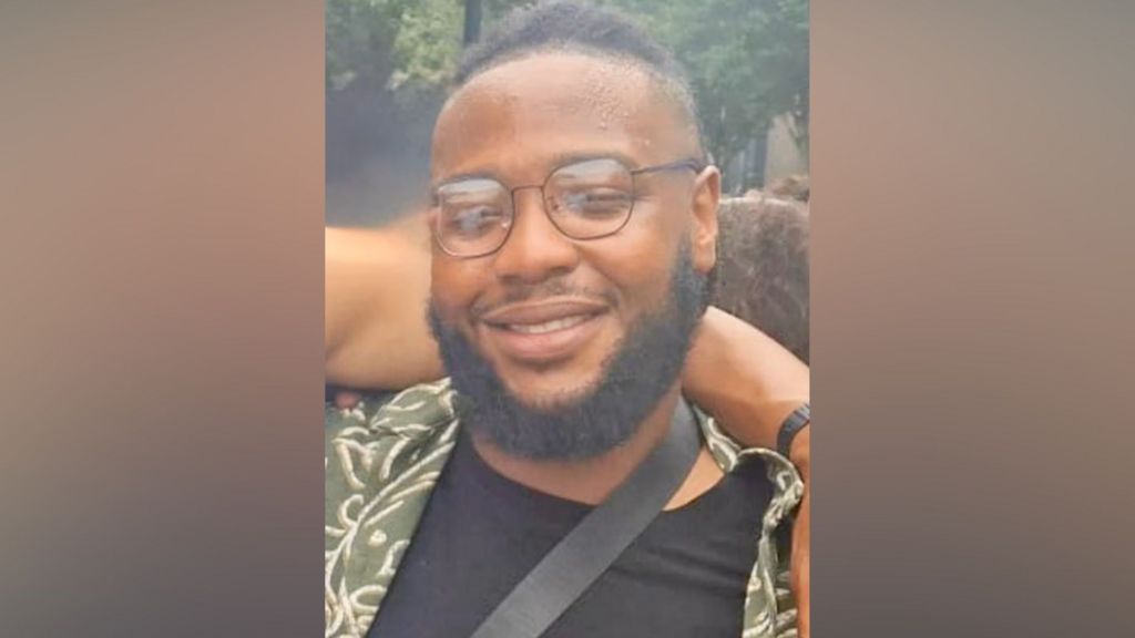 Close-up portrait of a smiling person with glasses and a beard, wearing a patterned shirt and black top, outdoors with trees in the background.