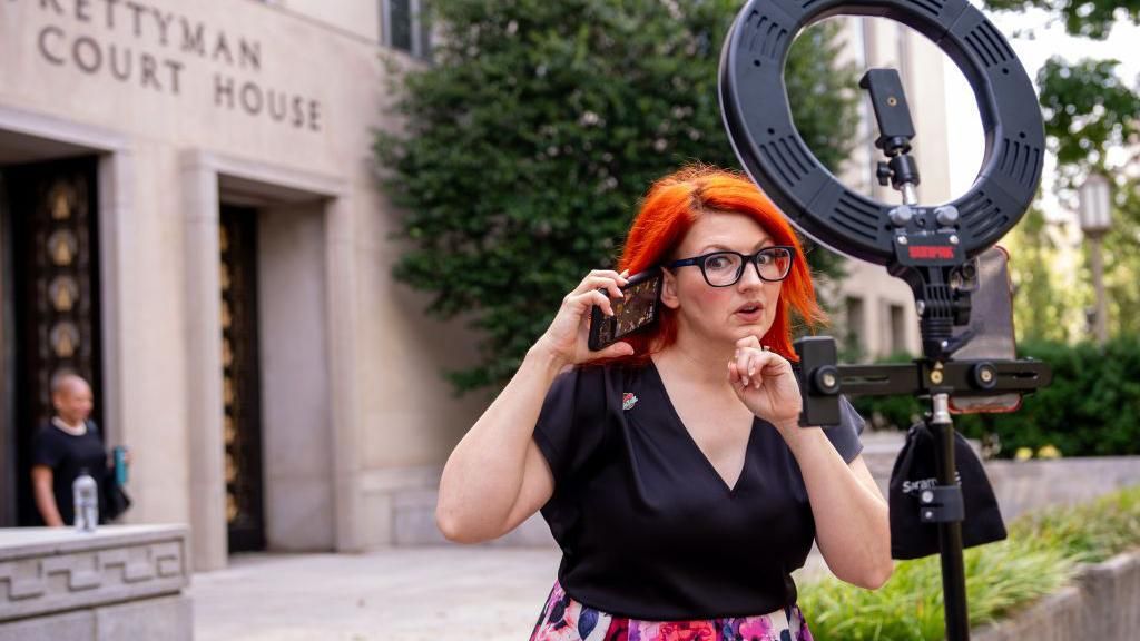 Tiffany Cianci shown holding her smartphone to her ear while filming herself on a smartphone attached to a ring light outside a courthouse in Washington, DC.