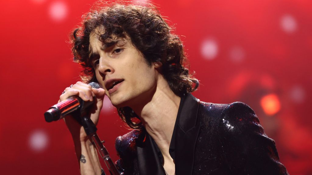 Sombr in a shiny dark jacket singing into a microphone on a leaning stand in front of a red background at the 2025 MTV Video Music Awards