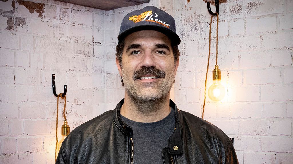 Rob Delaney wearing a hat and standing in front of a white brick wall