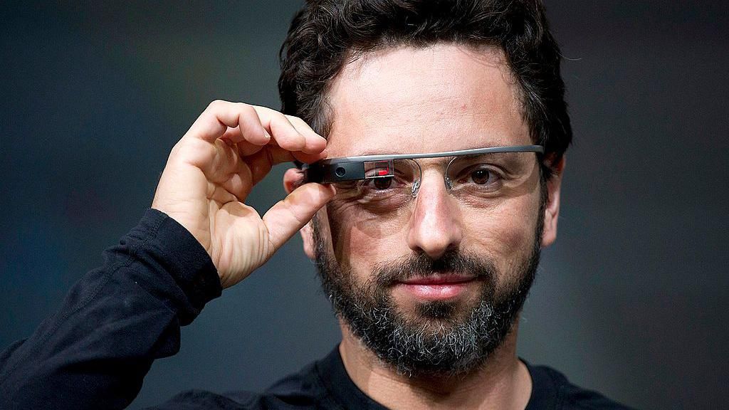 Sergey Brin wears a black long-sleeved top and Google Glass. He is looking directly into the lens of the camera capturing him, with one hand hovering over the right side of Google's digital glasses. On the right lens of the glasses, beside a black box containing the glasses camera, is a small red and black digital display.