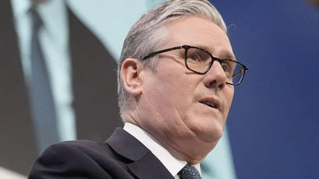 Prime Minister Starmer speaking at the conference. He is wearing a black suit and white shirt with a dark-coloured patterened tie.