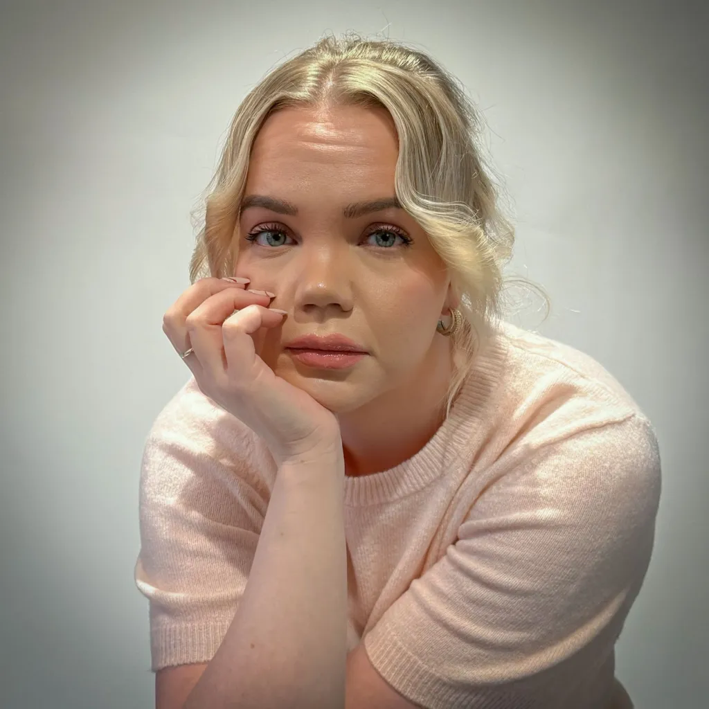 Meri-Tuuli Auer looking towards the camera, she is seated with her head resting in her hand. She has blonde hair and is wearing a light pink short sleeved woollen top.