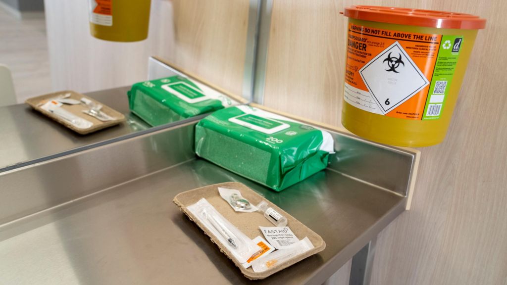 Inside an injection bay in the using space at the drug consumption room. On the counter is a brown cardboard tray with a number of items, including a syringe. There is also a packet of wipes and a yellow disposal bin fixed to the wall.