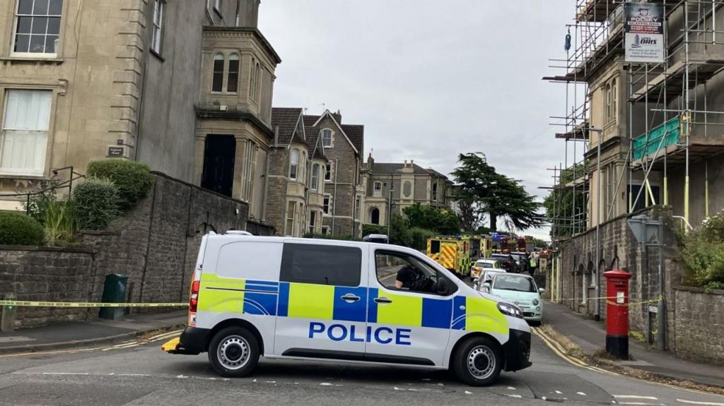 A police van blocking the entrance to Victoria Road. Police cars and ambulances are lined all up the street in the background.