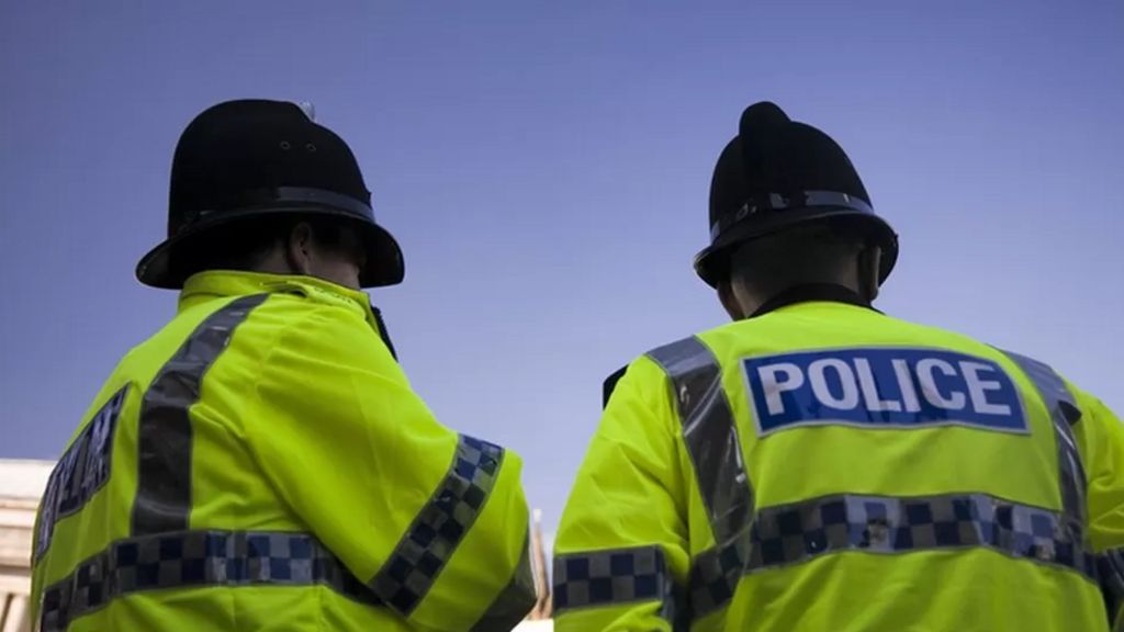 Two police officers in hi-vis jackets and black police hats stand together in front of a blue sky 