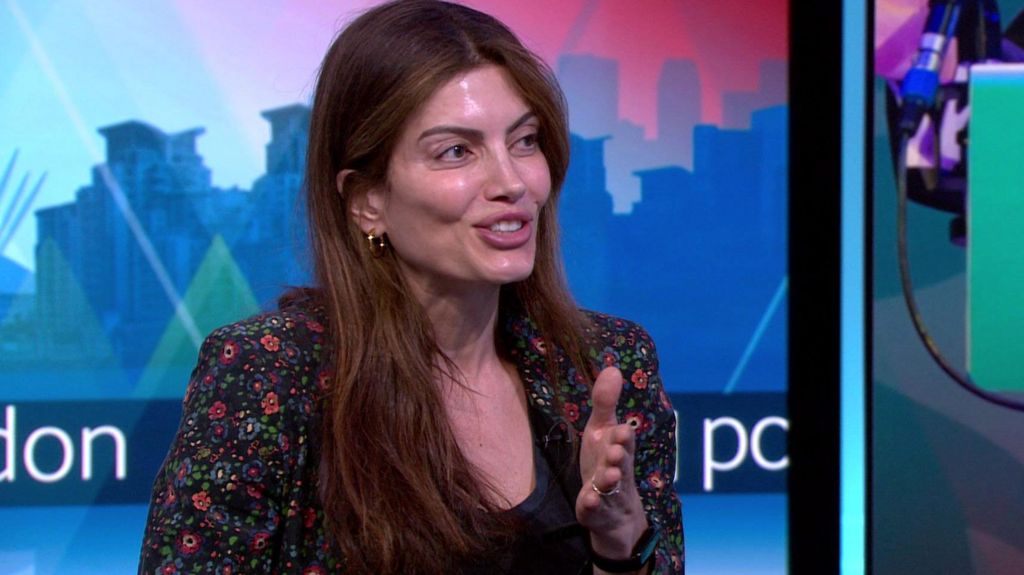 A woman in her thirties with long brown hair gestures as she is interviewed on a TV set. She is wearing a floral patterned jacket. 