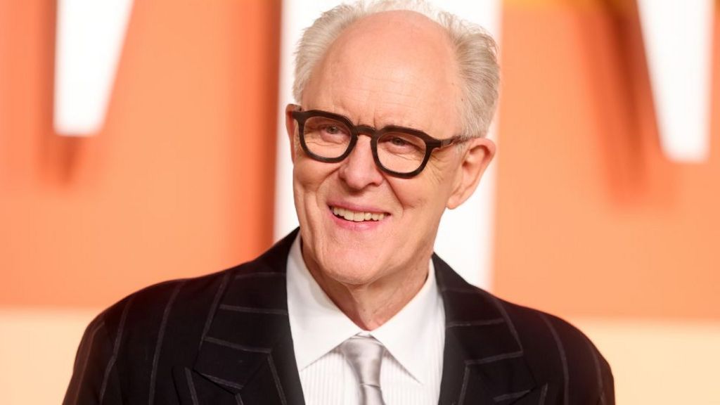 John Lithgow at the 2025 Vanity Fair Oscar Party. He is waring a black pinstriped jacket, white shirt and black-rimmed glasses.