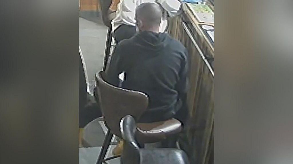 A man sitting on a bar stool with his back to the camera. He is wearing a black hoodie