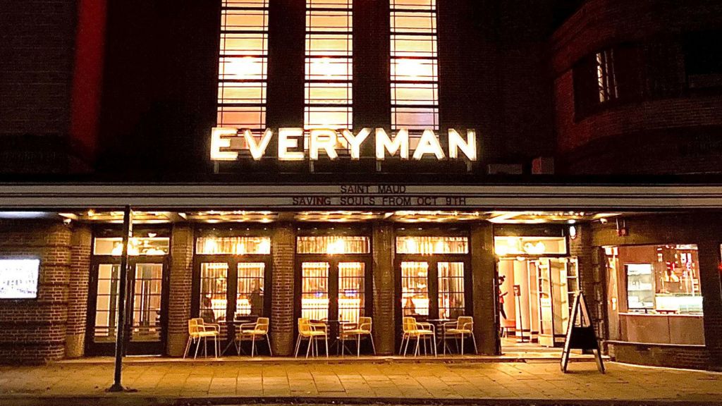 The facade of an art deco-style cinema building at night. There are five patio glass doors with tables and chairs in front. The far right is the entrance to which the doors are wide open and a person can be seen walking inside. Above the doors is a lit LED sign stating: EVERYMAN.
