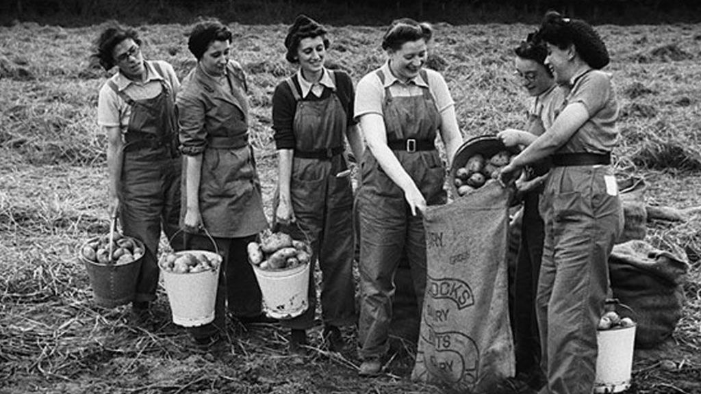 Six World War Two Land Army women wearing dungarees with buckets of potatoes