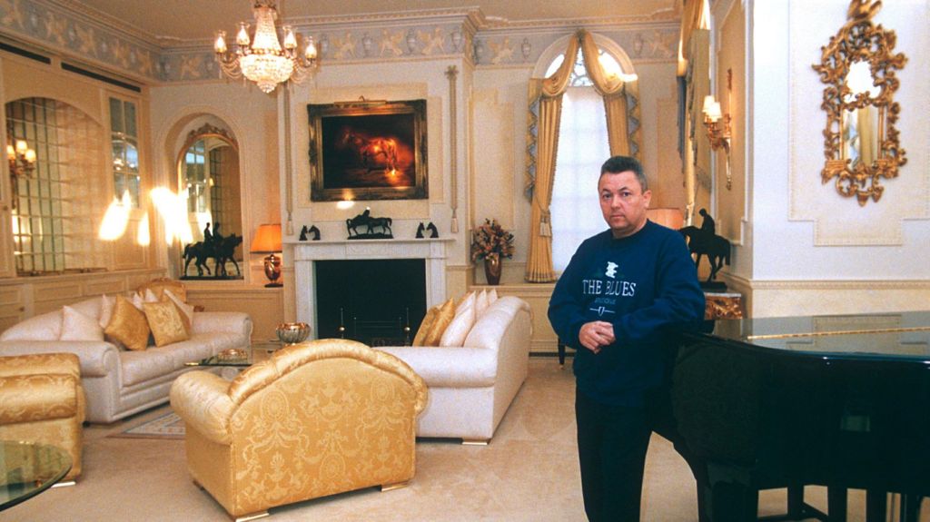 David Sullivan pictured at home in the 1990s, wearing a sweatshirt which says "The Blues" in reference to Birmingham City which he then co-owned. He is pictured leaning against a grand piano in an ornately decorated living room, with a crystal chandelier, gilt mouldings and mirrors, and sofas in white and gold.