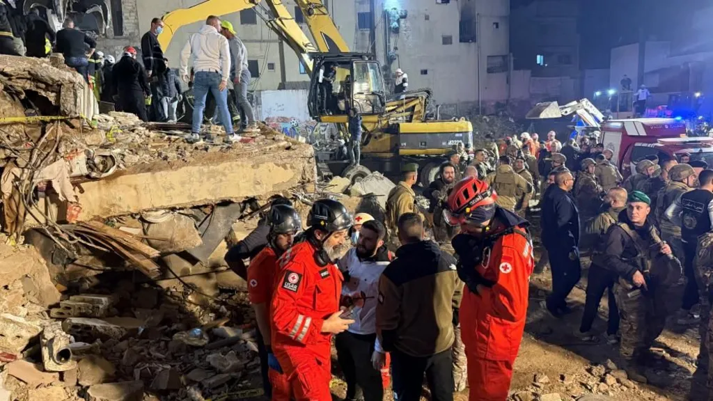 Emergency workers in red outfits standing at the site of collapsed buildings as an excavator is pictured in the background with a crowd of people gathered in and around piles of rubble, in Tripoli, Lebanon (08/02/26)