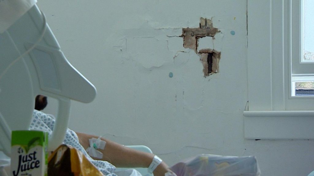 A man in a hospital bed, only his arm is visible. There is a hole in the wall behind him, through which plaster and lath is clearly visible.