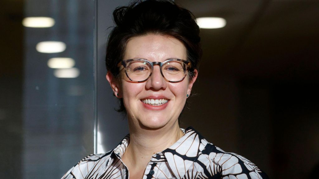 Claire Lynch smiling, wearing glasses and a black-and-white patterned shirt