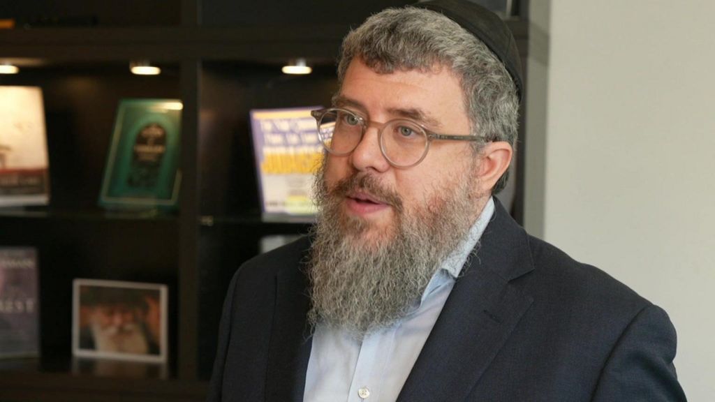 Rabbi Bentzi Sudak, wearing glasses, a black kippah, and a dark suit, speaks in front of a bookshelf.