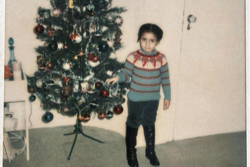 Five-year-old Andrea is wearing a bright festive jumper and standing next to a Christmas tree in what appears to be a living room.
