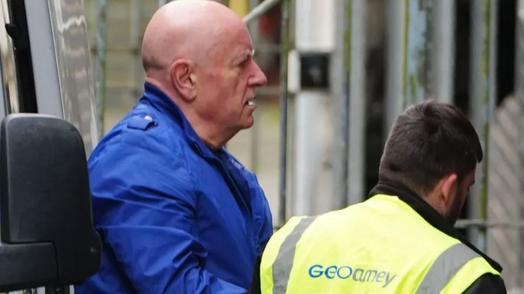 Foden, a bald man wearing a blue jacket, is led towards a policed van by a man with his back to the camera wearing yellow high viz