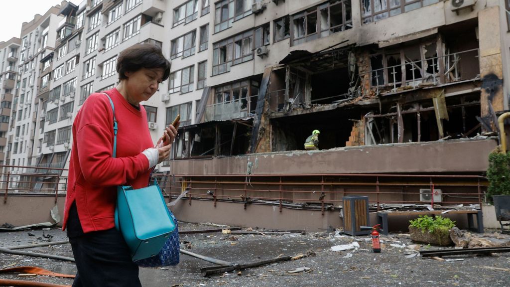 A woman wearing a red jumper and holding her phone walks past a bombed-out residential building in Kyiv on 21 July.