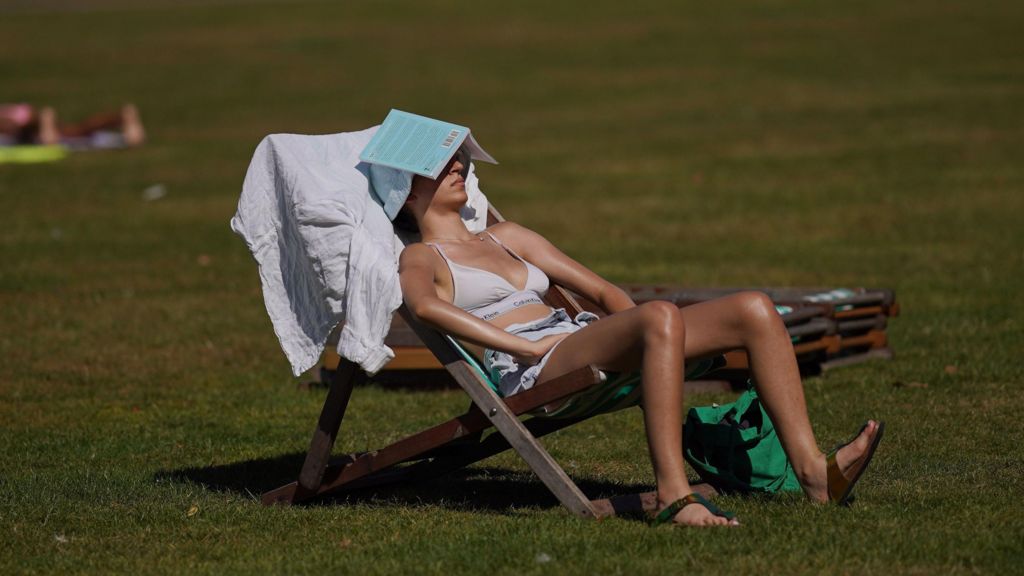 A woman sits on a deckchair on a sunny day in London. She is wearing a cream bra top and has a book resting on her head to shield her eyes from the sun.