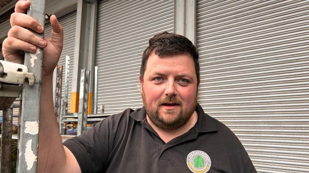 Plant Market owner Sean Thomas,who has short brown hair and a beard, is wearing a black T-shirt and stands in front of closed roller shutters in a warehouse