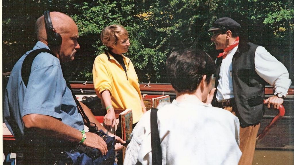Sir David Suchet wearing a black cap and waistcoat aboard a narrowboat with Pam Rhodes and camera crew
