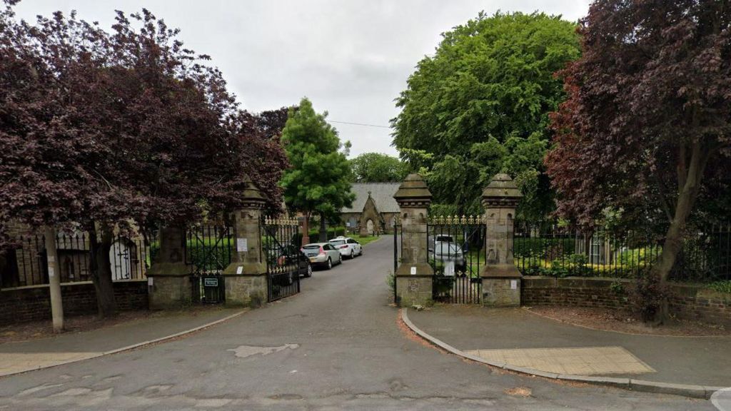 The entrance to Dean Road Cemetery in Scarborough, with trees either side of the gates and a road leading to the building.