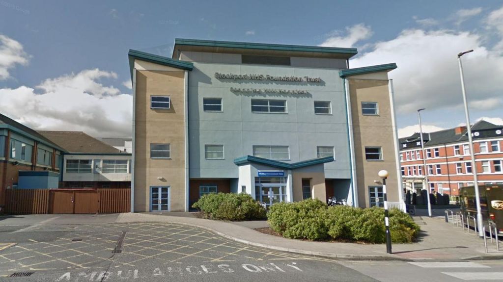 External view of Stepping Hill Hospital which is grey and beige. Ambulance bay at front of building with bushes outside of the entrance and a pelican crossing. 