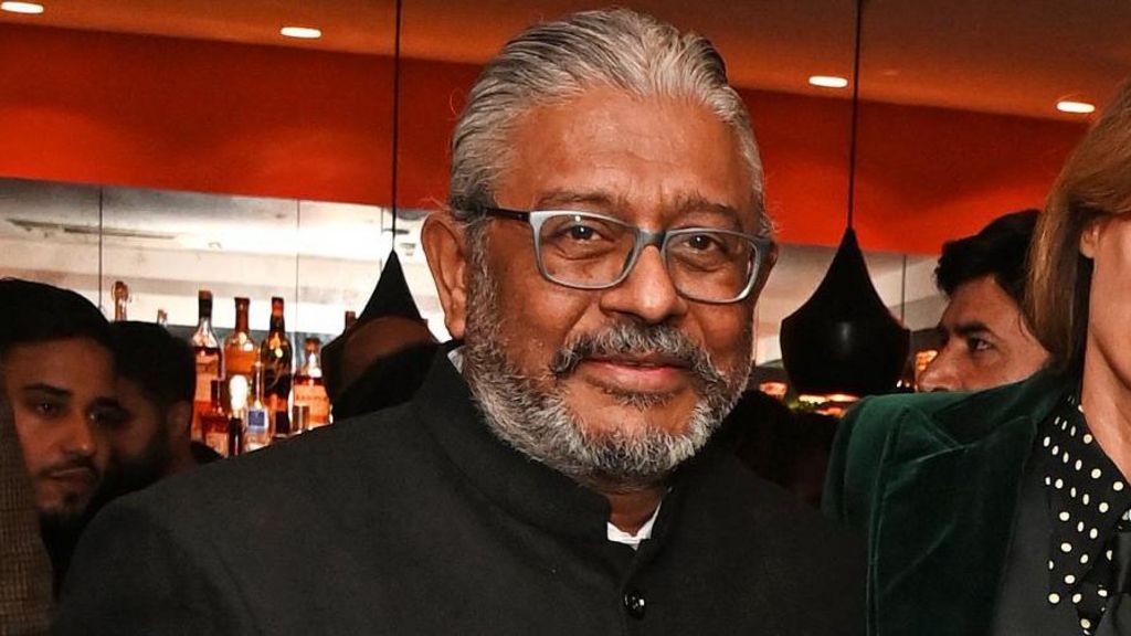 A middle-aged man with a short grey beard, wearing a high-necked suit jacket, looking at the camera.
